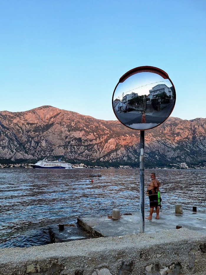 Road mirror reflecting the village, with the bay and mountains behind